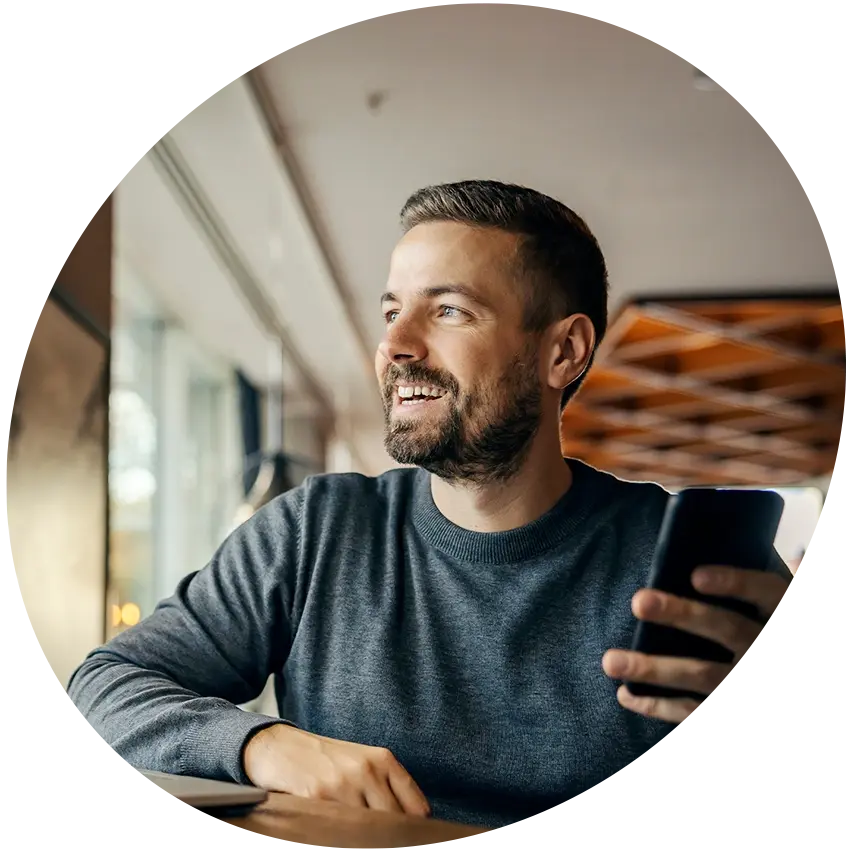 Young professional man working on a laptop while smiling at his phone, sitting on a sofa in a modern office lounge.