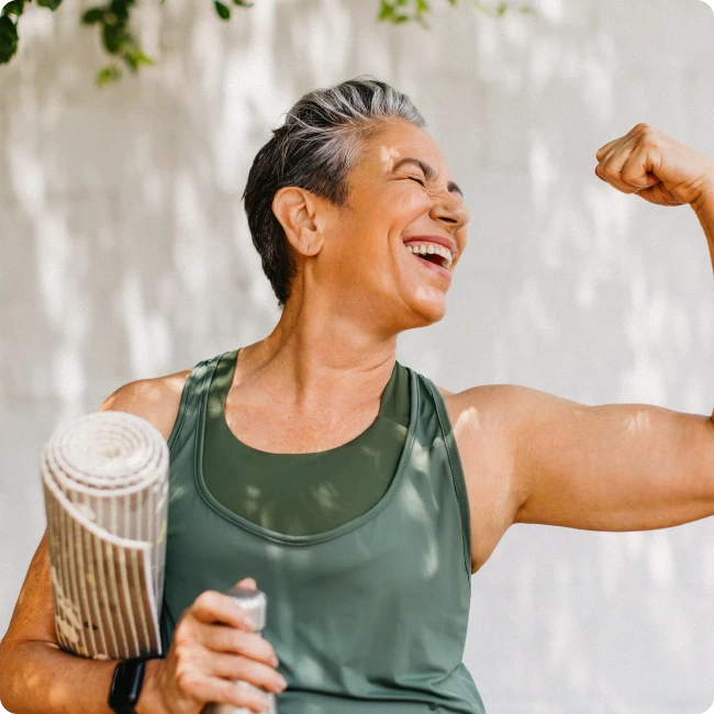 Older lady with a yoga mat flexing her muscles and smiling.